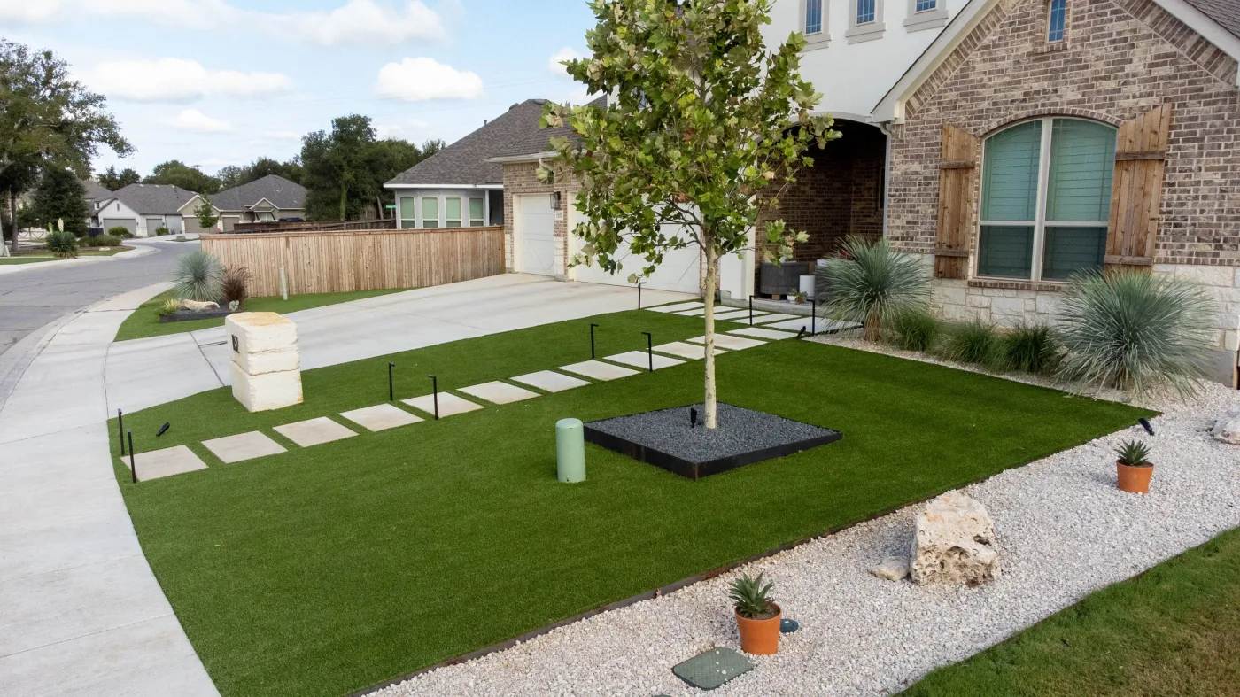 An artificial turf front lawn with square concrete steppers, a young shade tree, and agaves along a brick home