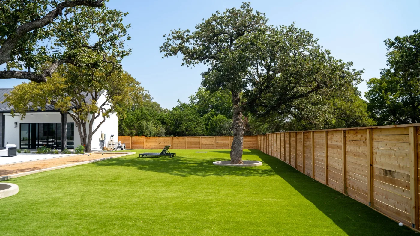 A large artificial turf backyard with a live oak set in a concrete tree ring, bordered by a new cedar fence