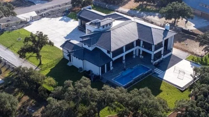 Aerial view of a large custom Hill Country home with a pool, sport court, and lawn among live oaks