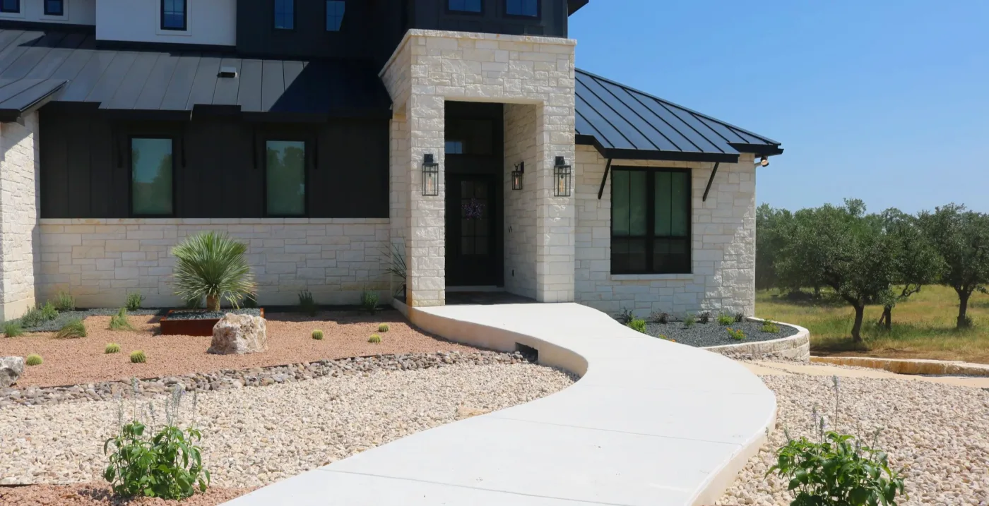 A limestone Hill Country home entry with a curving concrete walkway through gravel beds planted with yucca and cactus