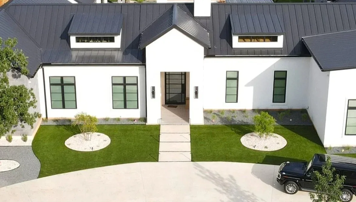 A white farmhouse front yard with a turf lawn, a stone walkway, and young trees ringed with light gravel