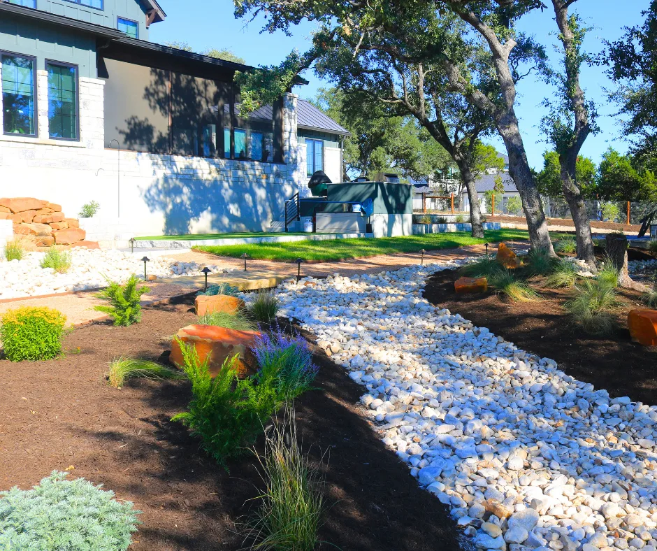 A dry creek bed of white river rock winding through mulched beds of native grasses and shrubs beside a Hill Country home