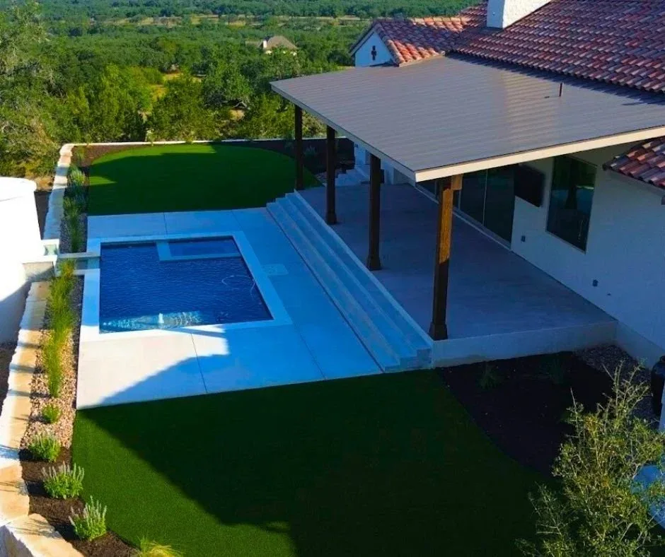 Aerial view of a backyard pool with artificial turf, a covered patio, and a limestone retaining wall overlooking open Hill Country