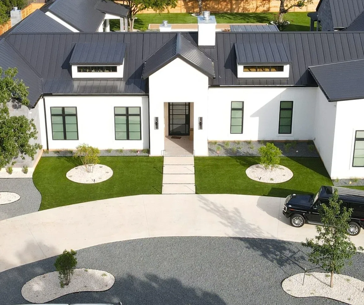 Aerial view of a modern white farmhouse with an artificial turf front lawn and gravel planting circles around young trees