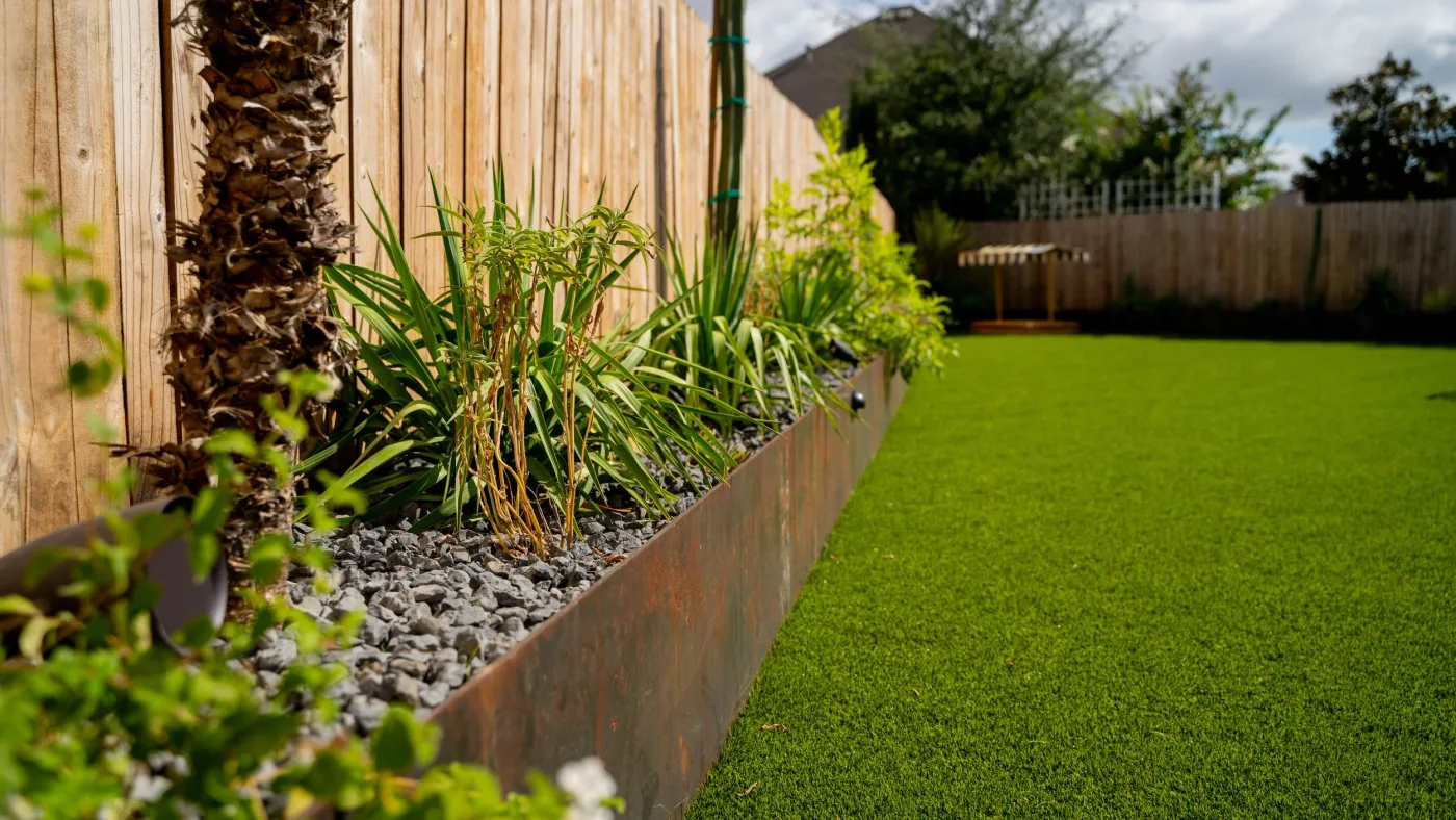 A steel-edged planting bed of grasses over gray gravel running along a cedar fence beside an artificial turf lawn