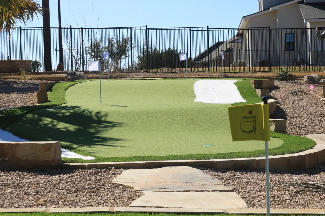 A backyard putting green with a flag and a sand bunker, edged in stone