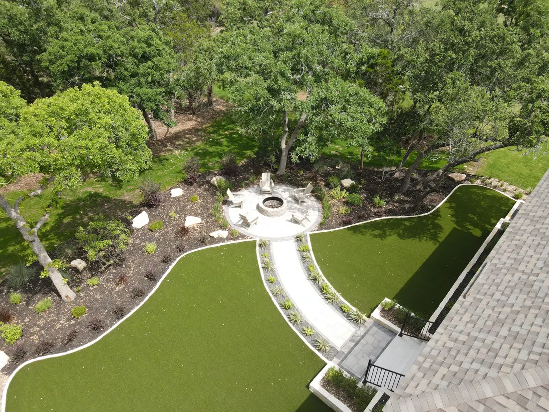 Aerial view of a landscape design installed across a whole backyard, with a turf lawn, a circular fire pit, and a paver path under mature oaks