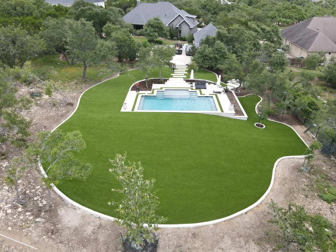 Aerial view of an artificial turf lawn wrapping a pool and spa on a wooded lot