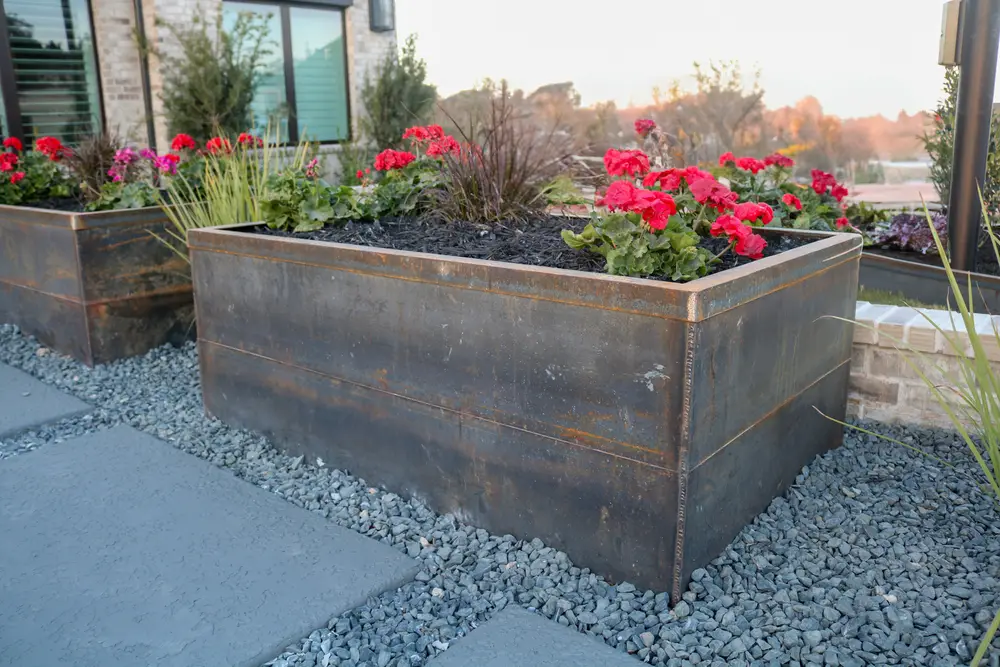 Corten steel planter filled with red geraniums and ornamental grasses over black gravel