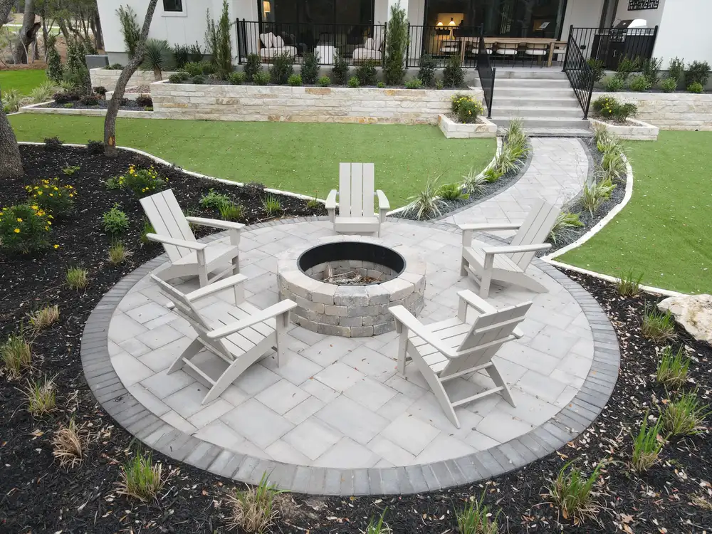 A circular paver patio with a fire pit and Adirondack chairs, set into a turf lawn