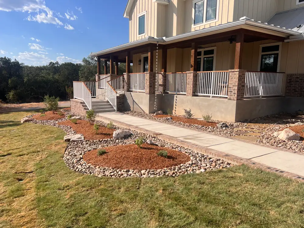 A wraparound farmhouse porch with a brick edged walk through river rock and mulch beds